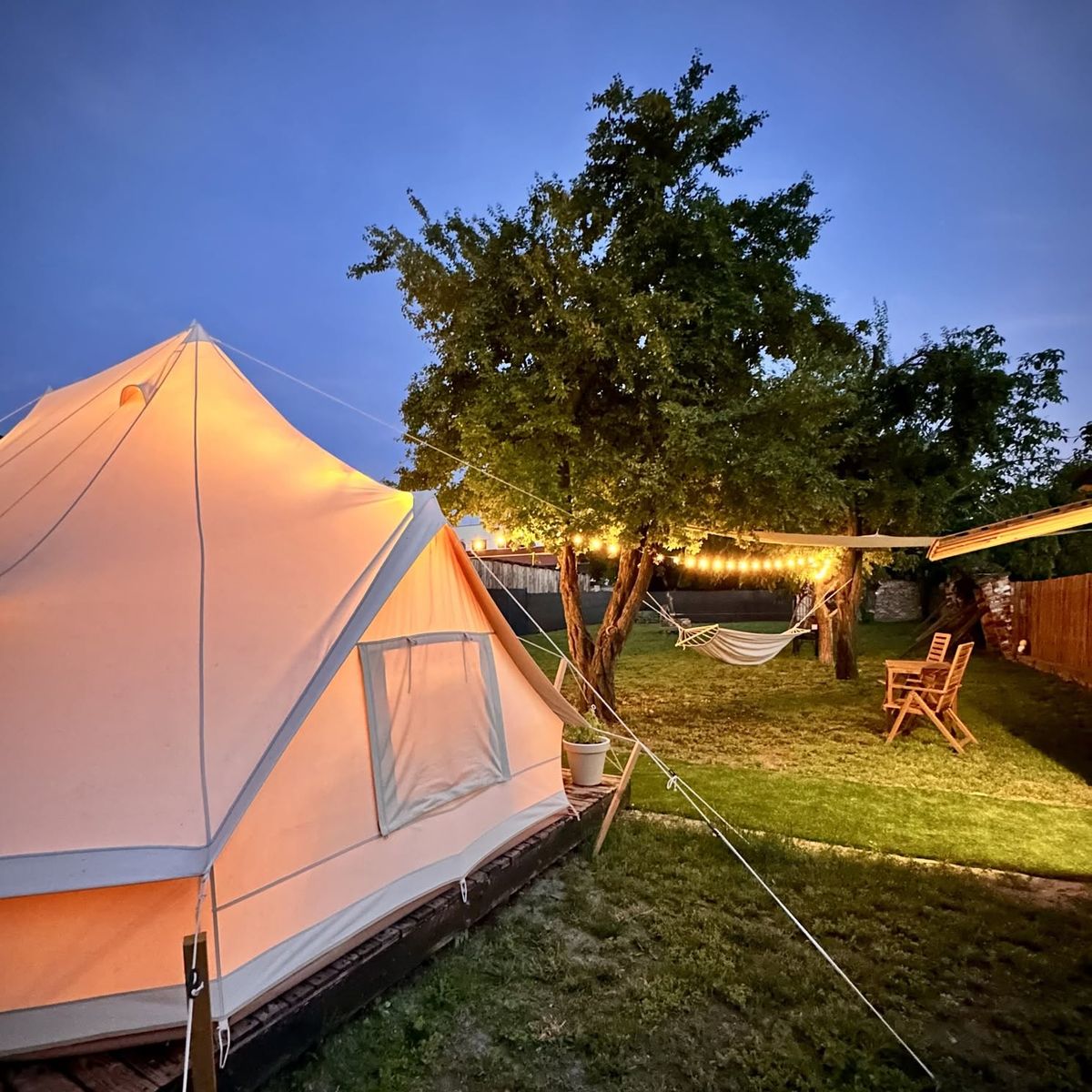Stan na dřevěné podlaze za soumraku, za ním hamak a židle pod rozsvícenými girlandami